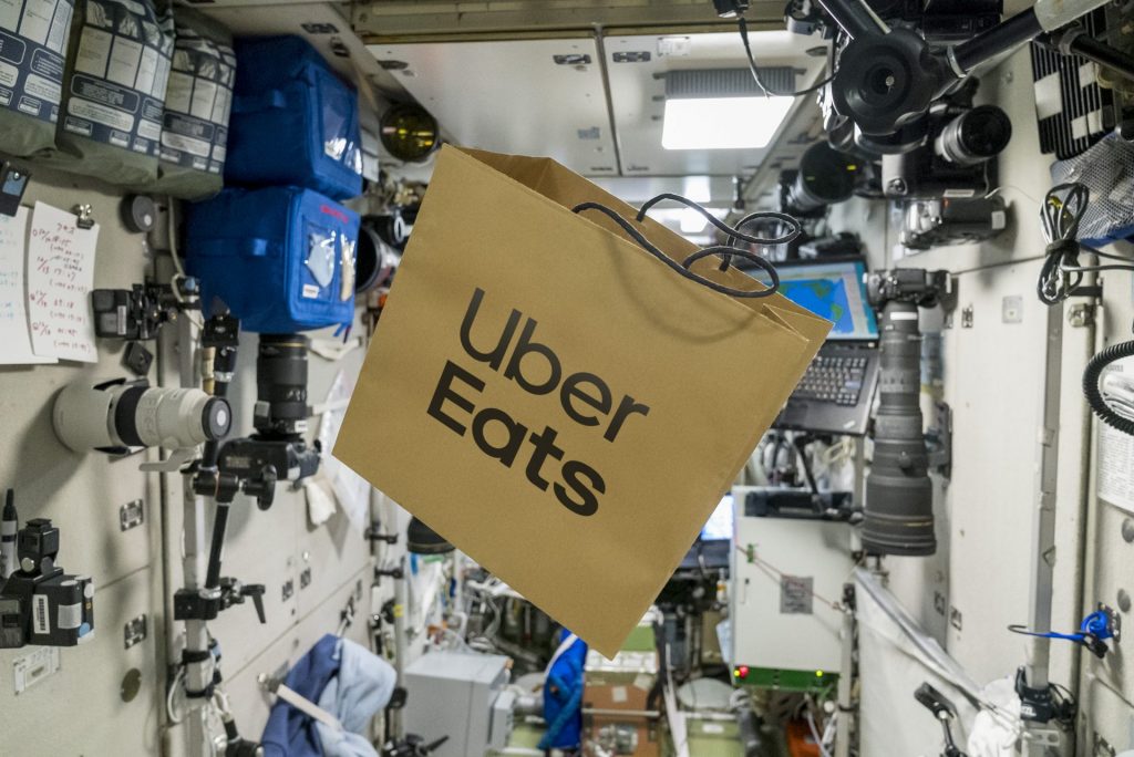 Imagem de uma sacola de papel com os dizeres Uber Eats impressos, pairando no ar dentro da nave russa, na estação espacial
