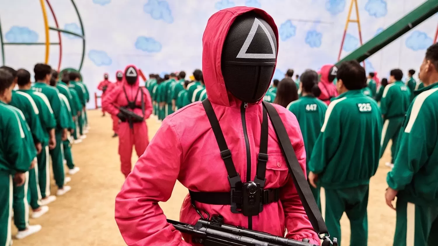 Trecho da série Round 6 da Netflix, com uma pessoa vestida com um macacão vermelho, segurando uma arma, e ao fundo uma fila de pessoas de macacão verde