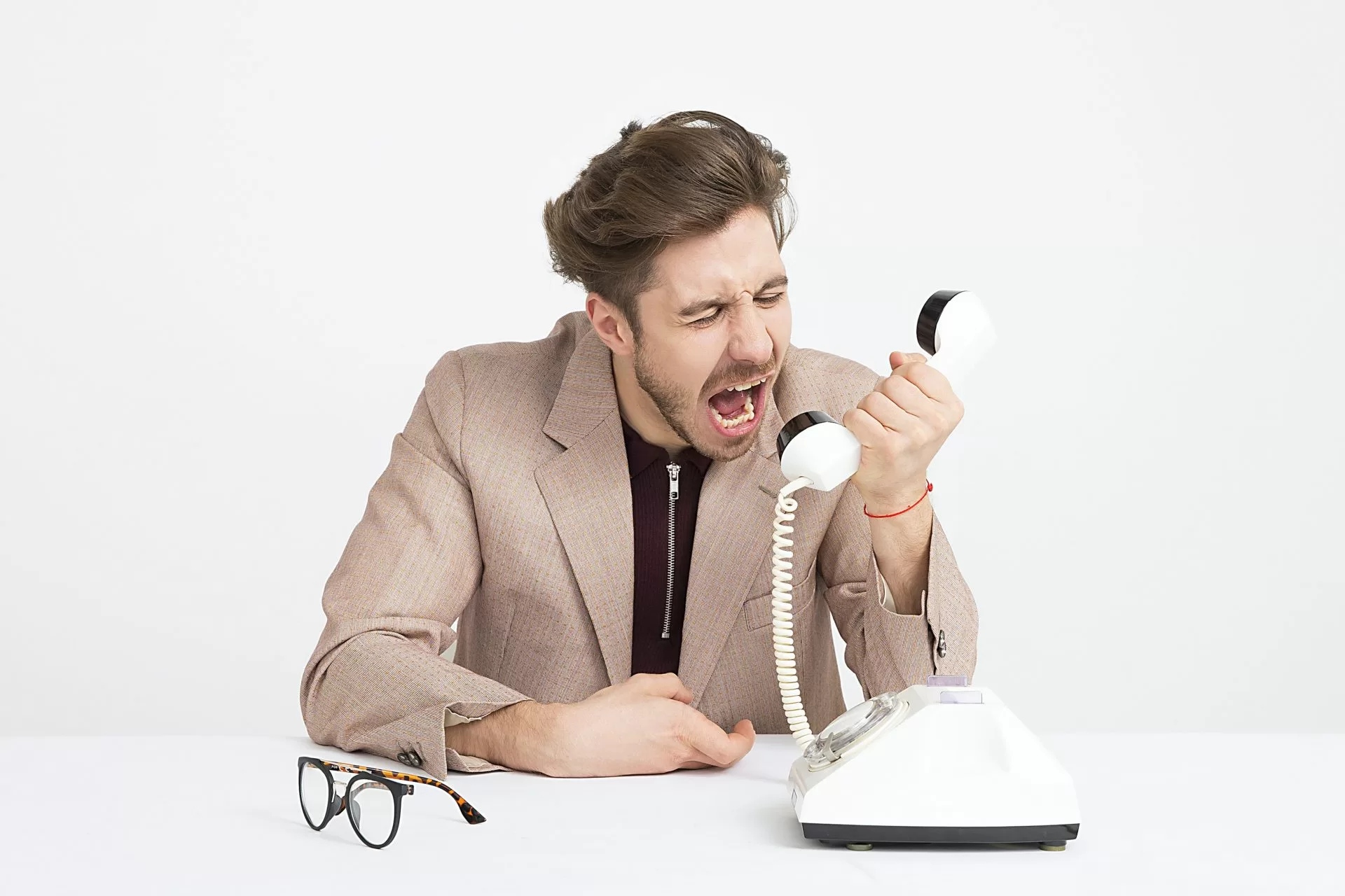Homem vestido com terno e sentado em uma mesa aparece gritando para um telefone fixo, como recebendo uma ligação indesejada de telemarketing