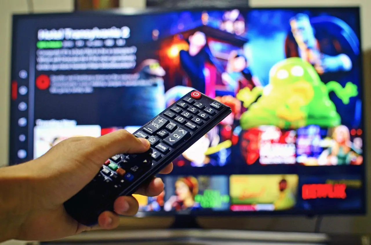 Imagem mostra uma mão segurando um controle remoto e, ao fundo, uma televisão com a tela inicial da Netflix sendo exibida